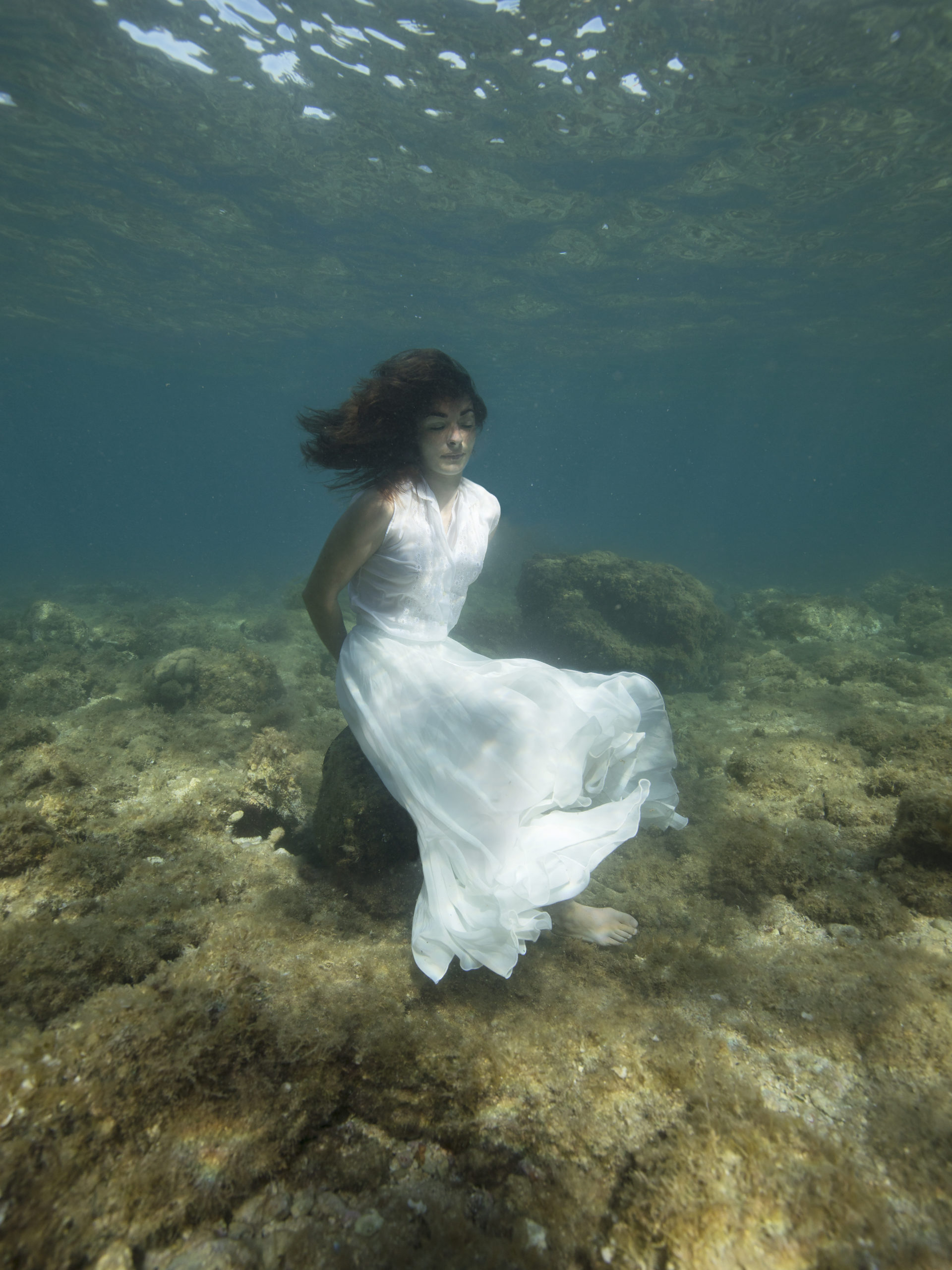 femme en robe blanche assise sur un banc de coquillage au fond de la mer.